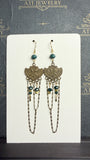 Long chandelier earrings with teal and golden-yellow beads, antique brass-tone bird plaques, and layered chain loops on a cream display card.