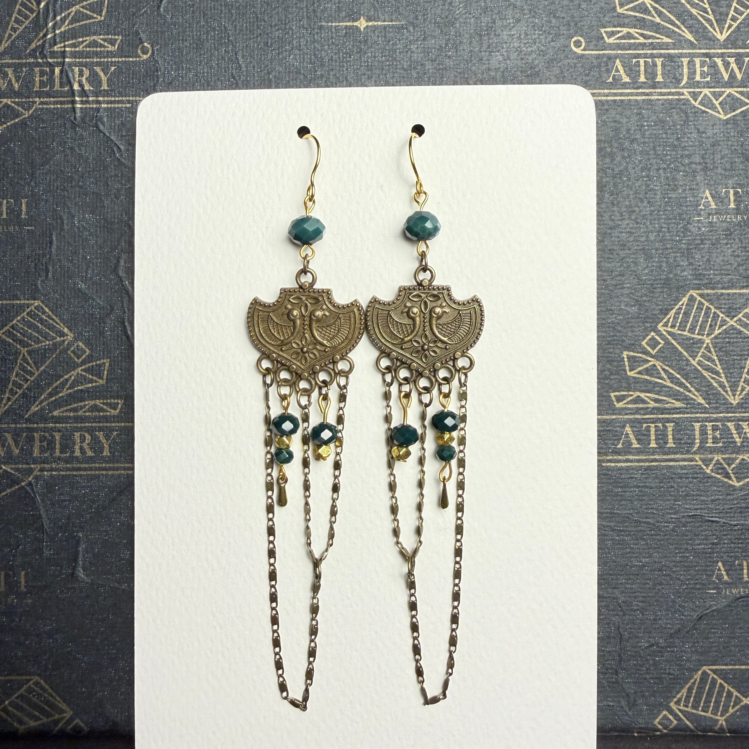 Long chandelier earrings with teal and golden-yellow beads, antique brass-tone bird plaques, and layered chain loops on a cream display card.