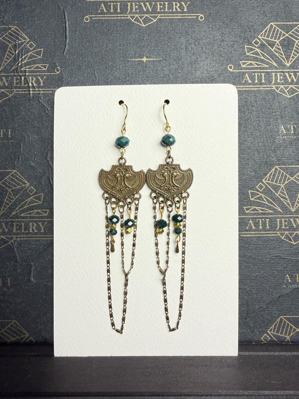 Long chandelier earrings with teal and golden-yellow beads, antique brass-tone bird plaques, and layered chain loops on a cream display card.