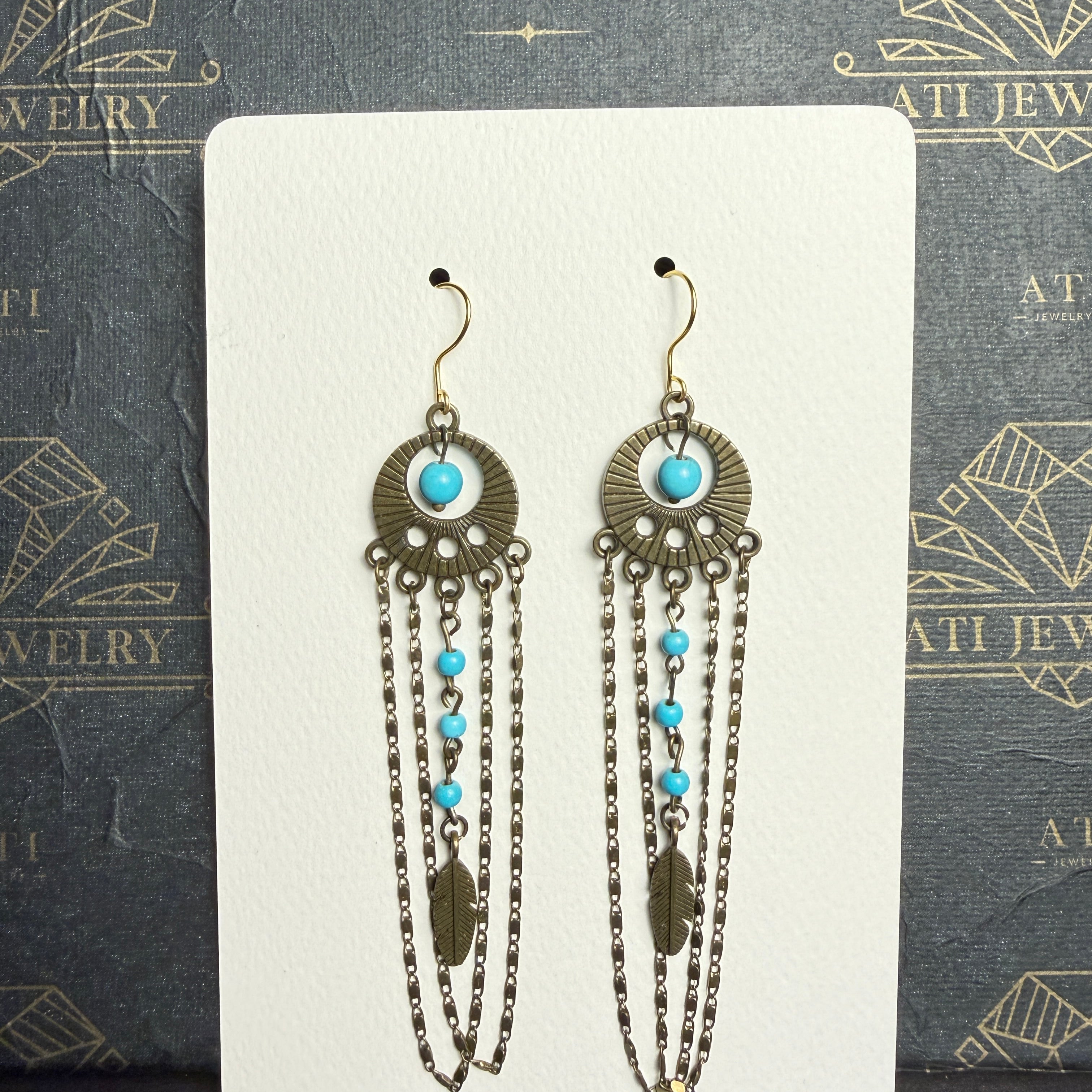 Long chandelier earrings with turquoise beads, antique brass-tone circular ornaments, feather charms, and layered chain loops on a cream display card.