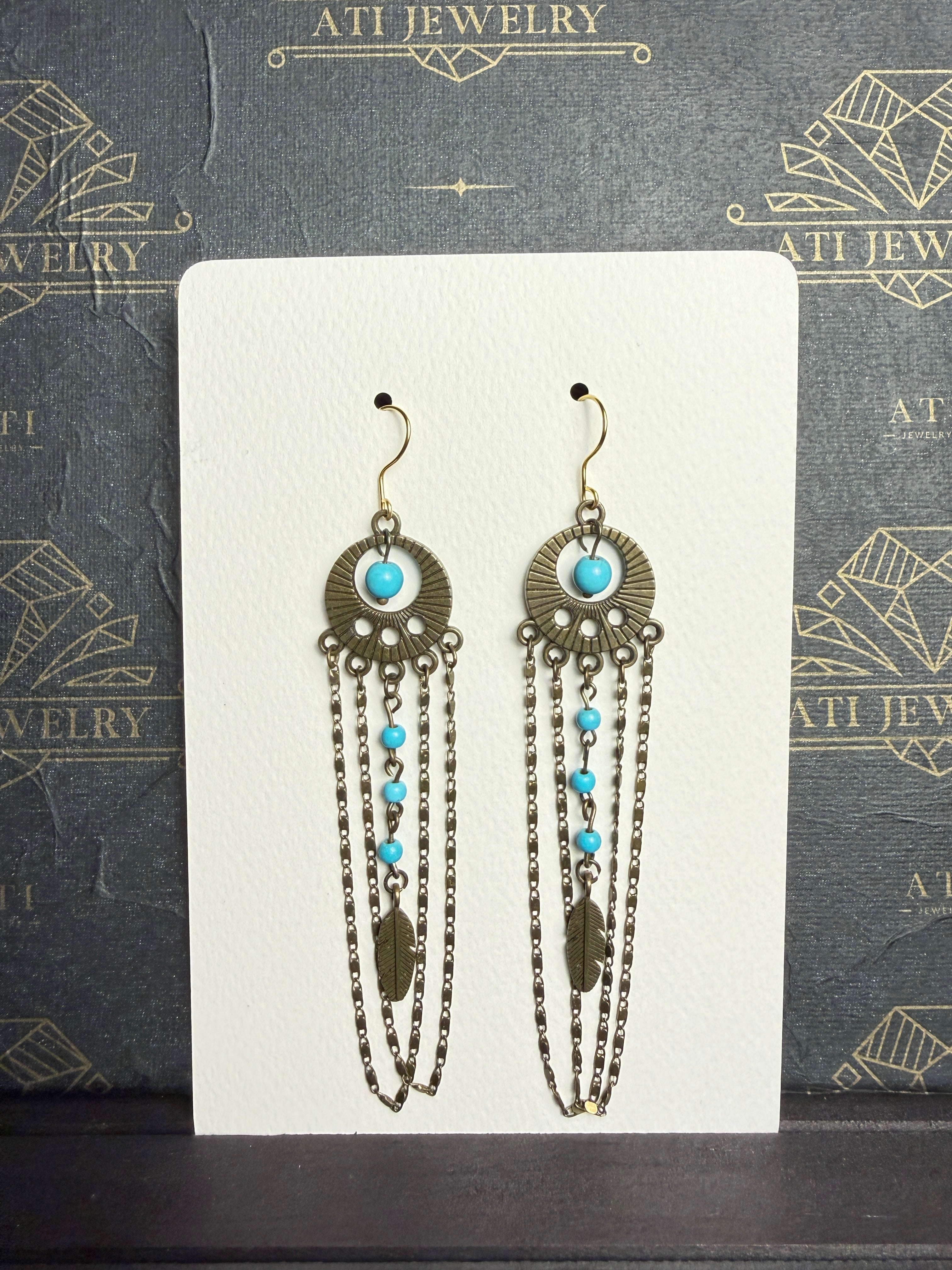 Long chandelier earrings with turquoise beads, antique brass-tone circular ornaments, feather charms, and layered chain loops on a cream display card.