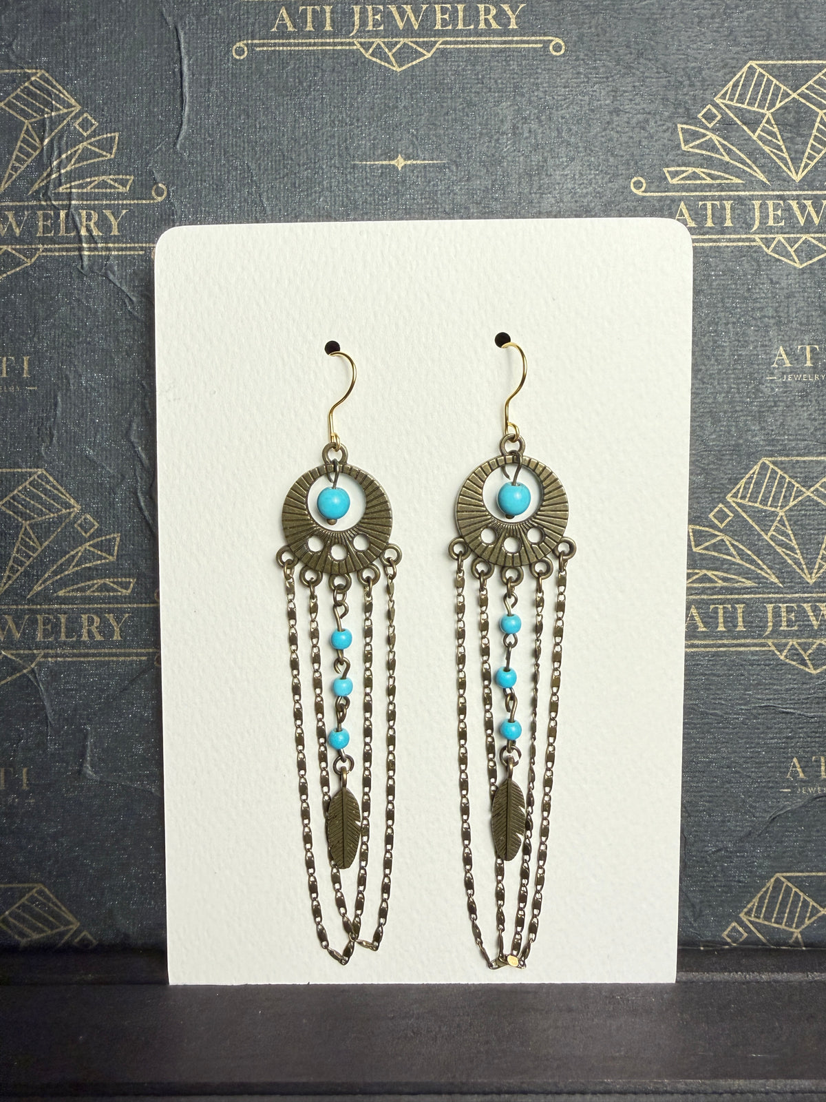 Long chandelier earrings with turquoise beads, antique brass-tone circular ornaments, feather charms, and layered chain loops on a cream display card.