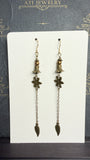 Long antique brass-tone earrings with bronze-coloured beads, bell-shaped accents, textured flowers, and chain drops ending in leaf charms.