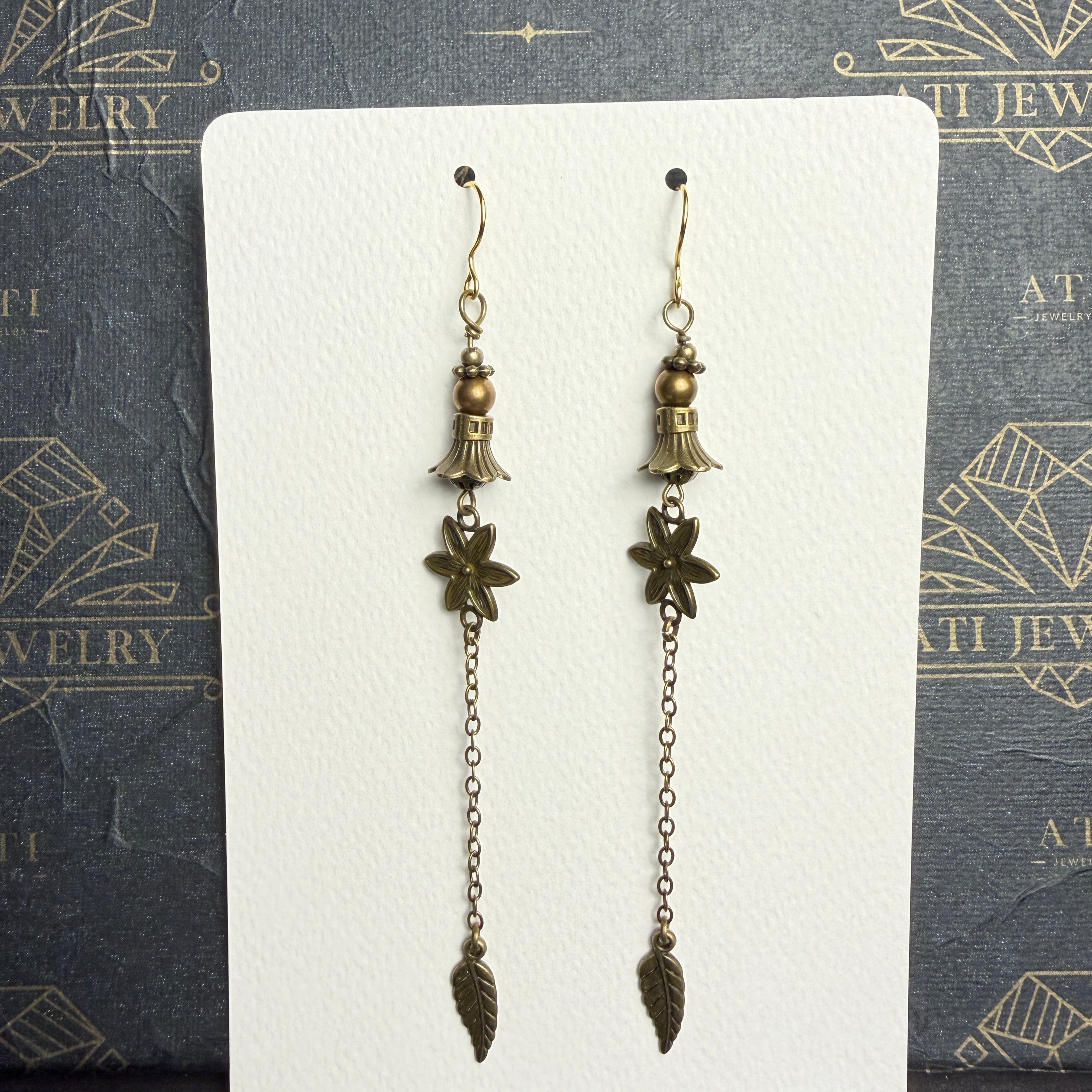 Long antique brass-tone earrings with bronze-coloured beads, bell-shaped accents, textured flowers, and chain drops ending in leaf charms.