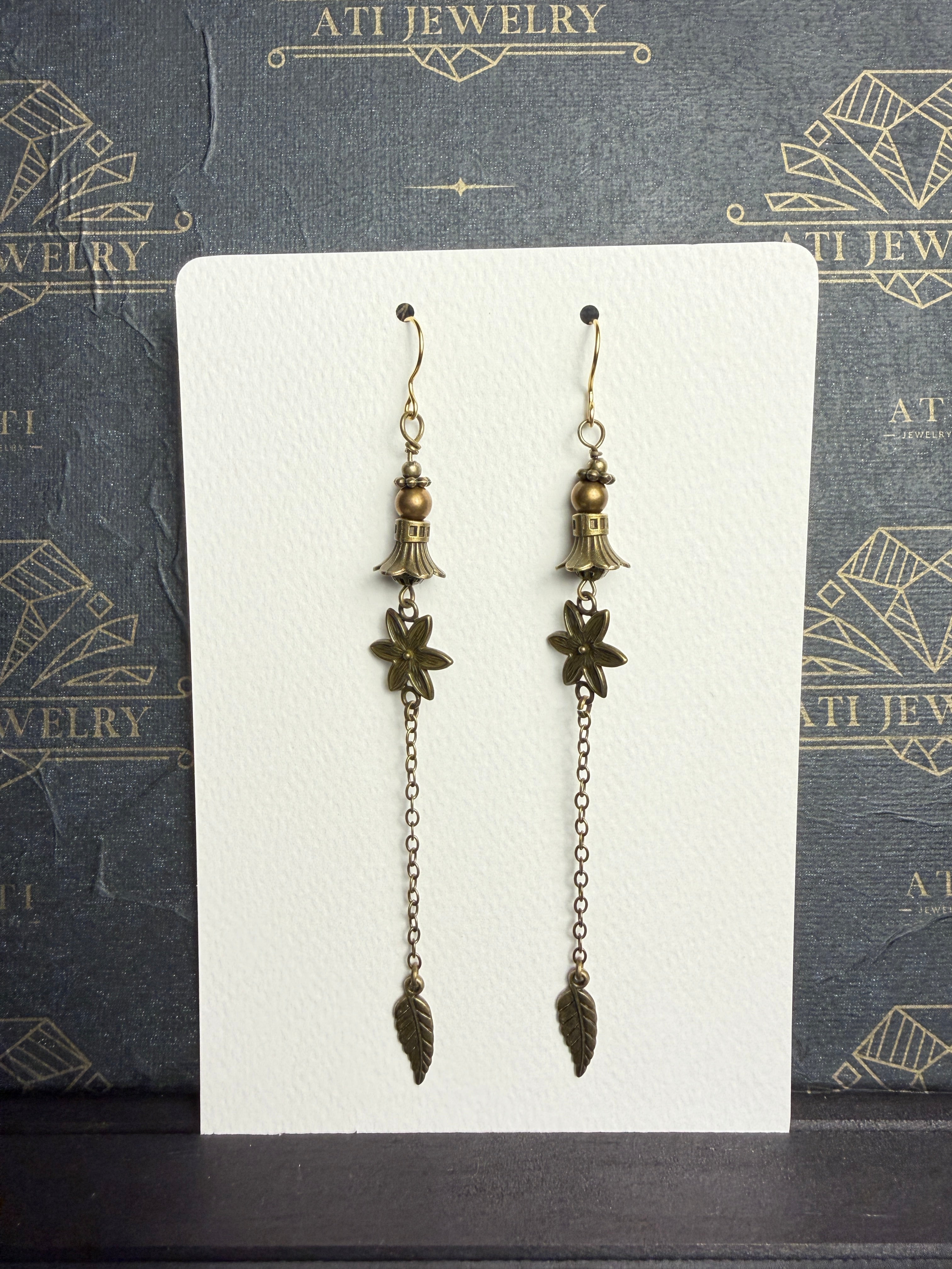 Long antique brass-tone earrings with bronze-coloured beads, bell-shaped accents, textured flowers, and chain drops ending in leaf charms.