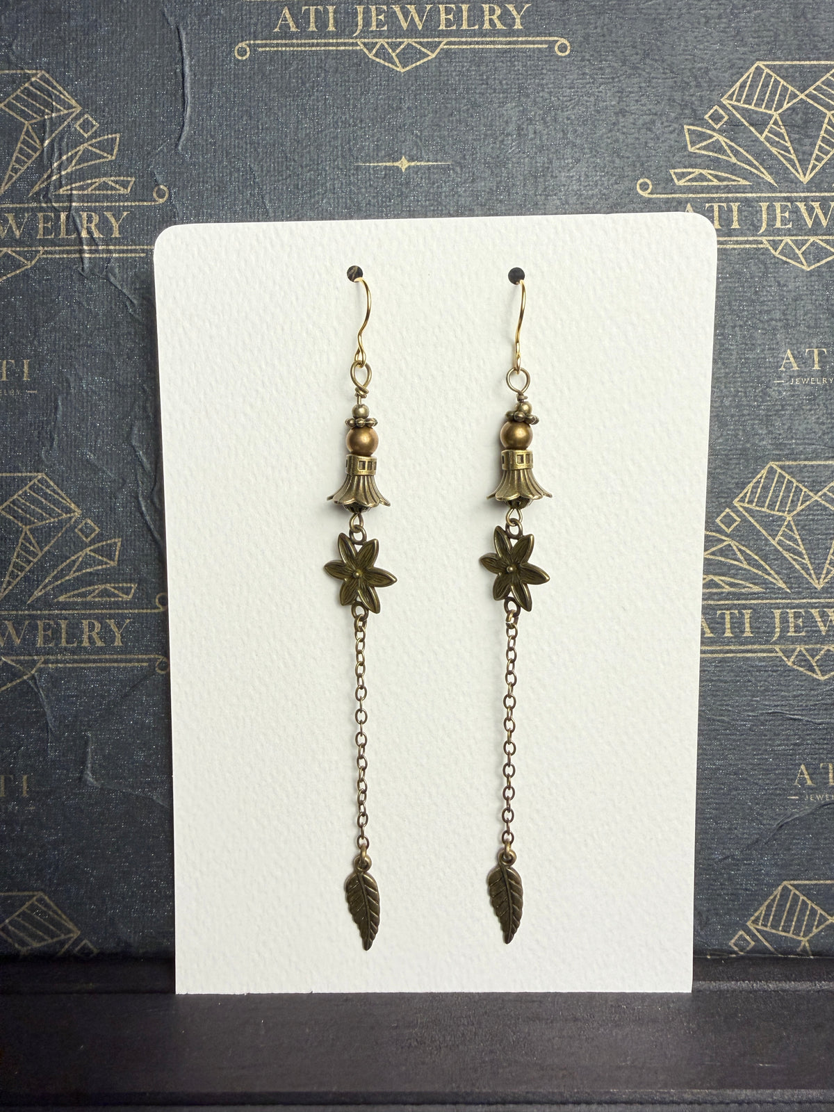 Long antique brass-tone earrings with bronze-coloured beads, bell-shaped accents, textured flowers, and chain drops ending in leaf charms.