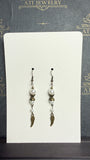 Long drop earrings with ivory pearl-like beads, ornate antique brass-tone spacers, and textured wing charms on an ATI JEWELRY display card.