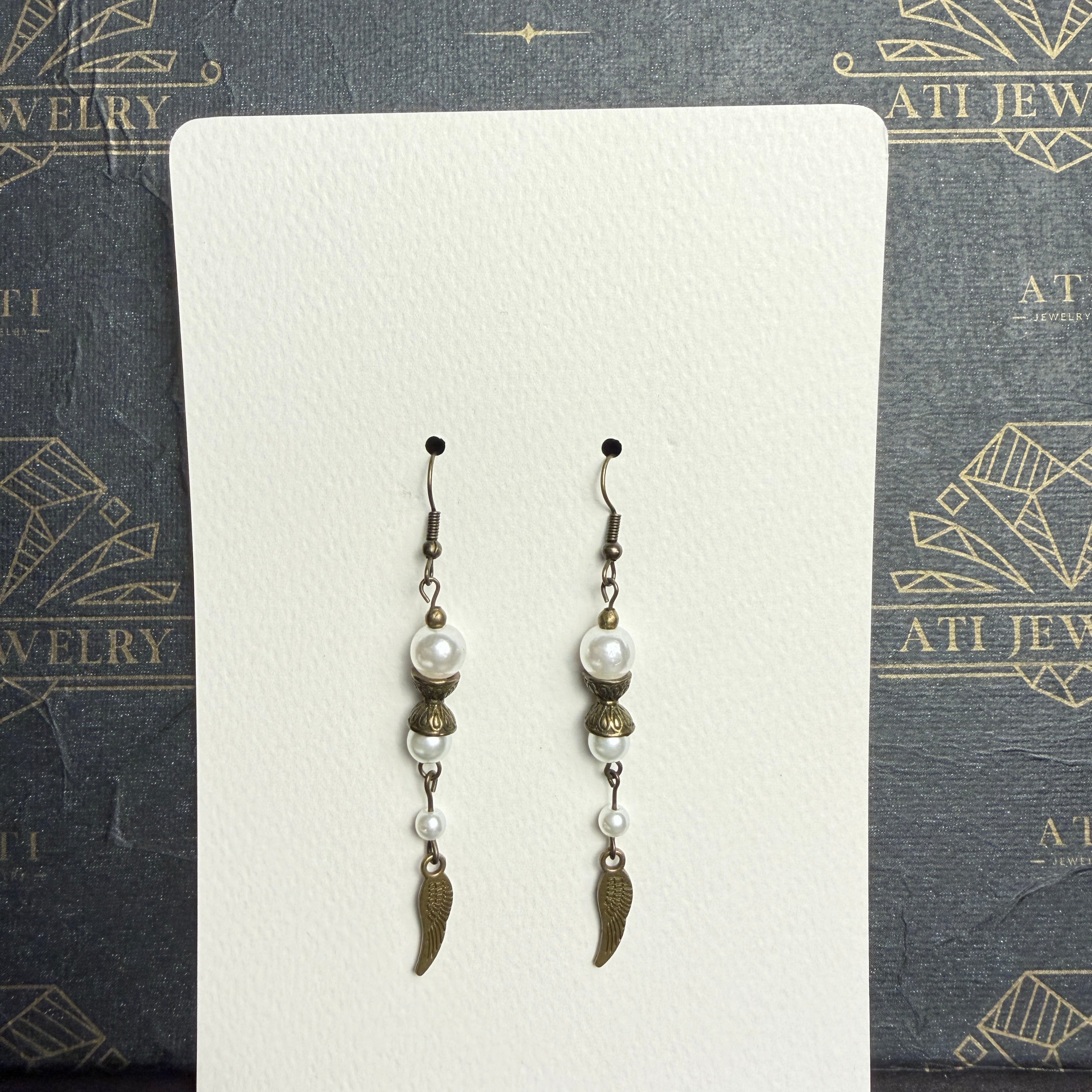Long drop earrings with ivory pearl-like beads, ornate antique brass-tone spacers, and textured wing charms on an ATI JEWELRY display card.