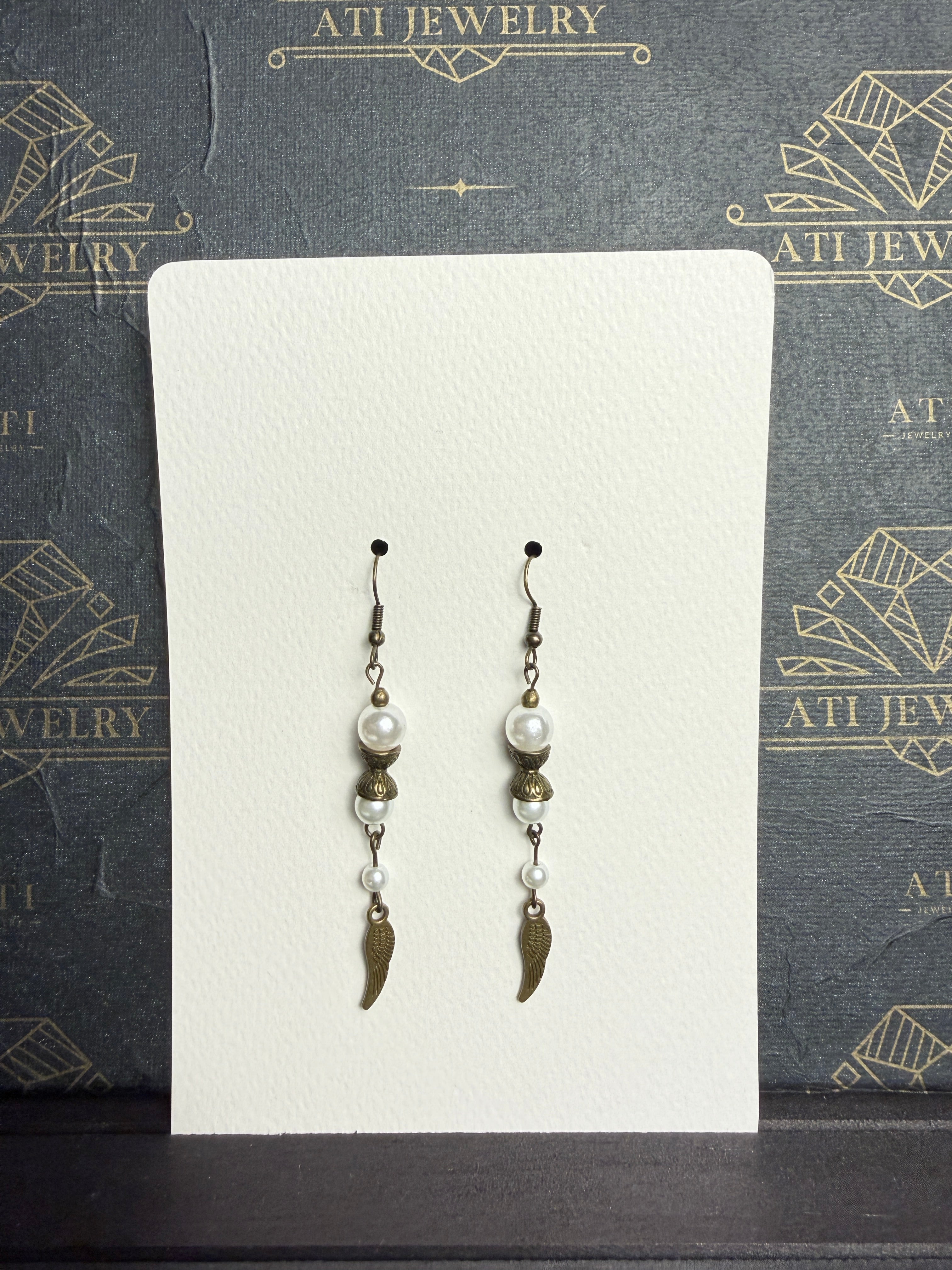 Long drop earrings with ivory pearl-like beads, ornate antique brass-tone spacers, and textured wing charms on an ATI JEWELRY display card.