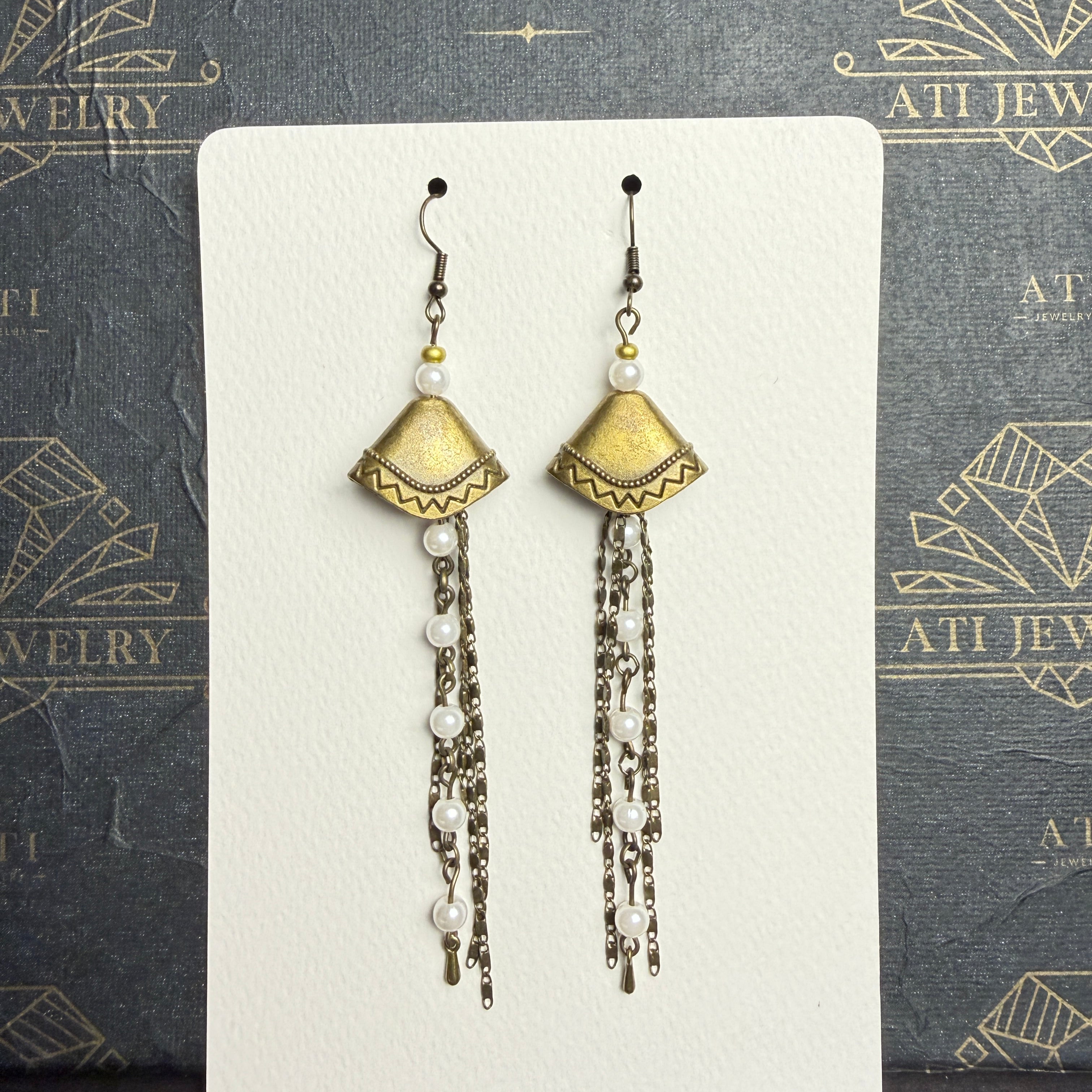 Long antique brass-tone earrings with geometric fan-shaped panels, white pearl-like beads, and layered chain tassels.