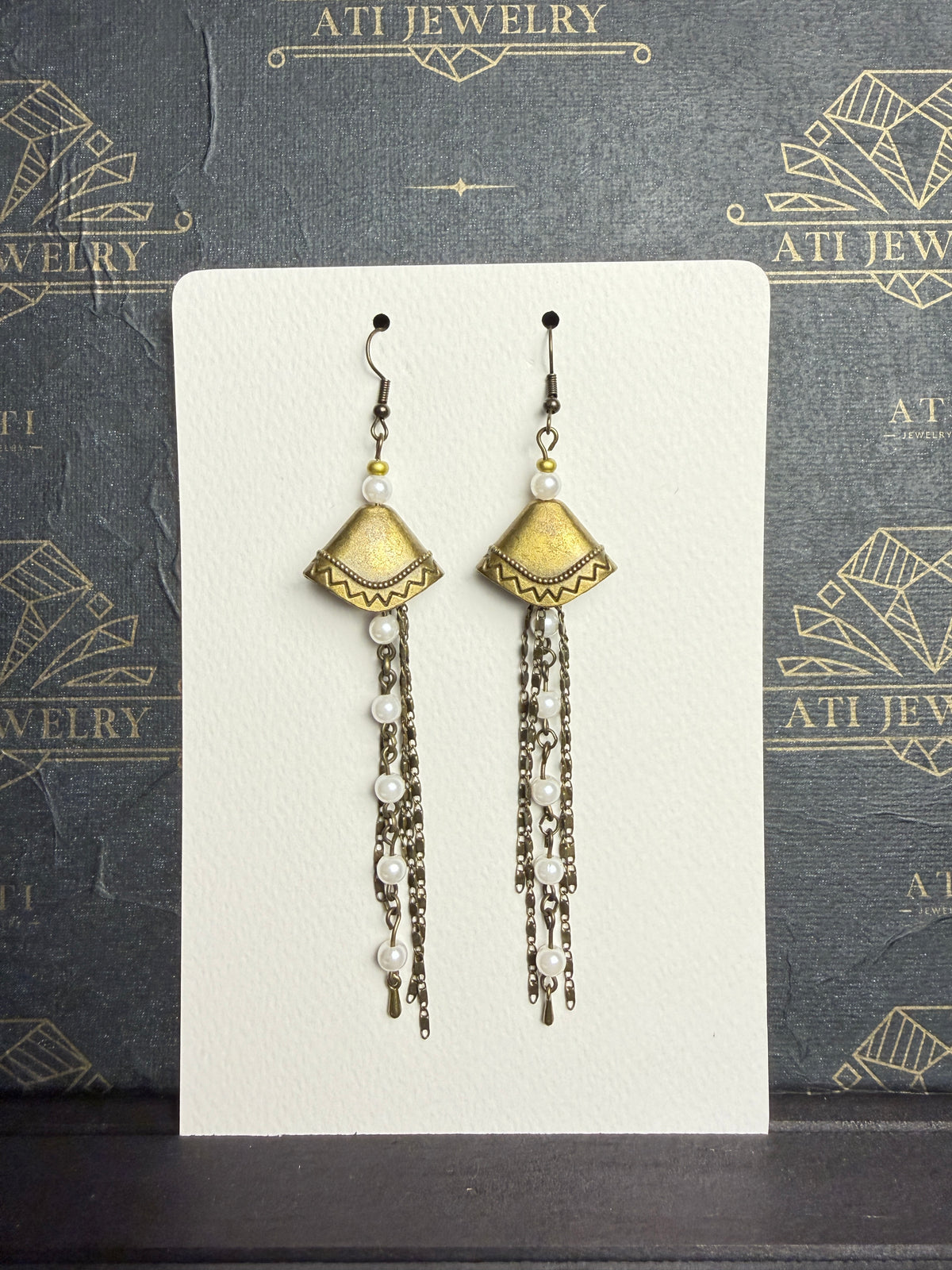 Long antique brass-tone earrings with geometric fan-shaped panels, white pearl-like beads, and layered chain tassels.