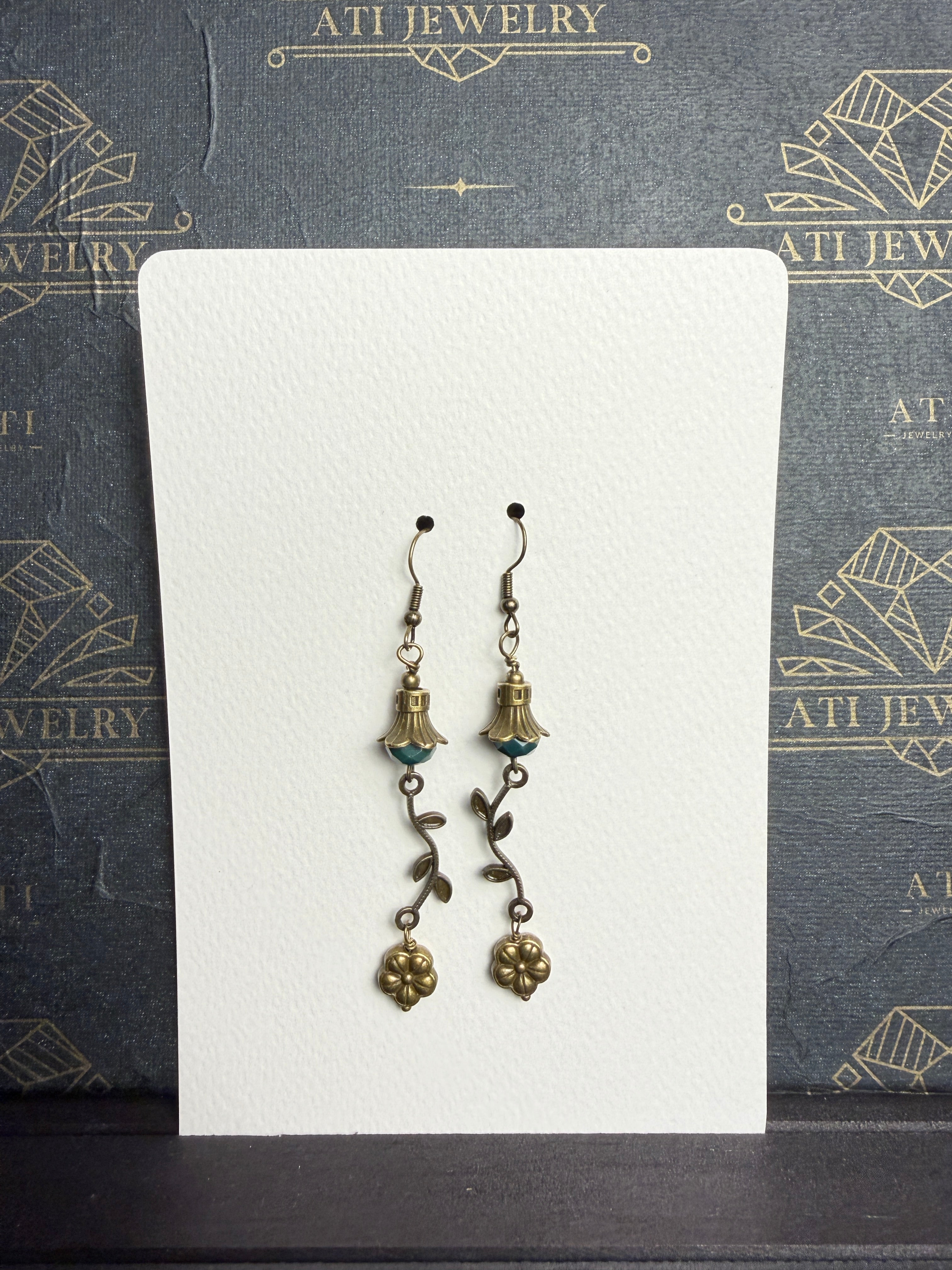 Antique brass-tone drop earrings with faceted teal beads, petal-shaped caps, curved leafy vines, and dangling flower charms.