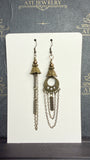 Mismatched antique brass-tone earrings with bell-shaped floral details, long chains, and an open circular ornament on one earring, on an ATI JEWELRY card.