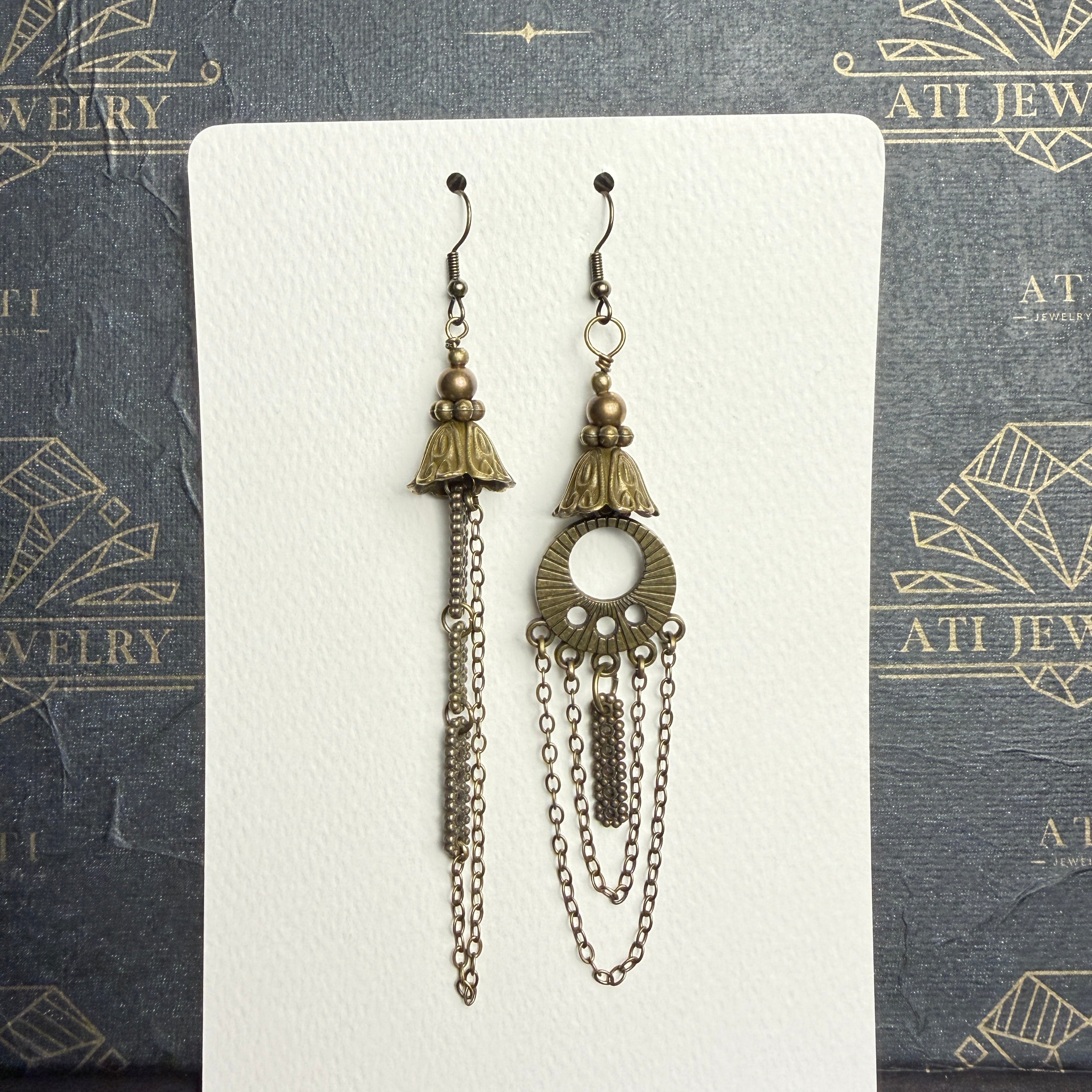 Mismatched antique brass-tone earrings with bell-shaped floral details, long chains, and an open circular ornament on one earring, on an ATI JEWELRY card.