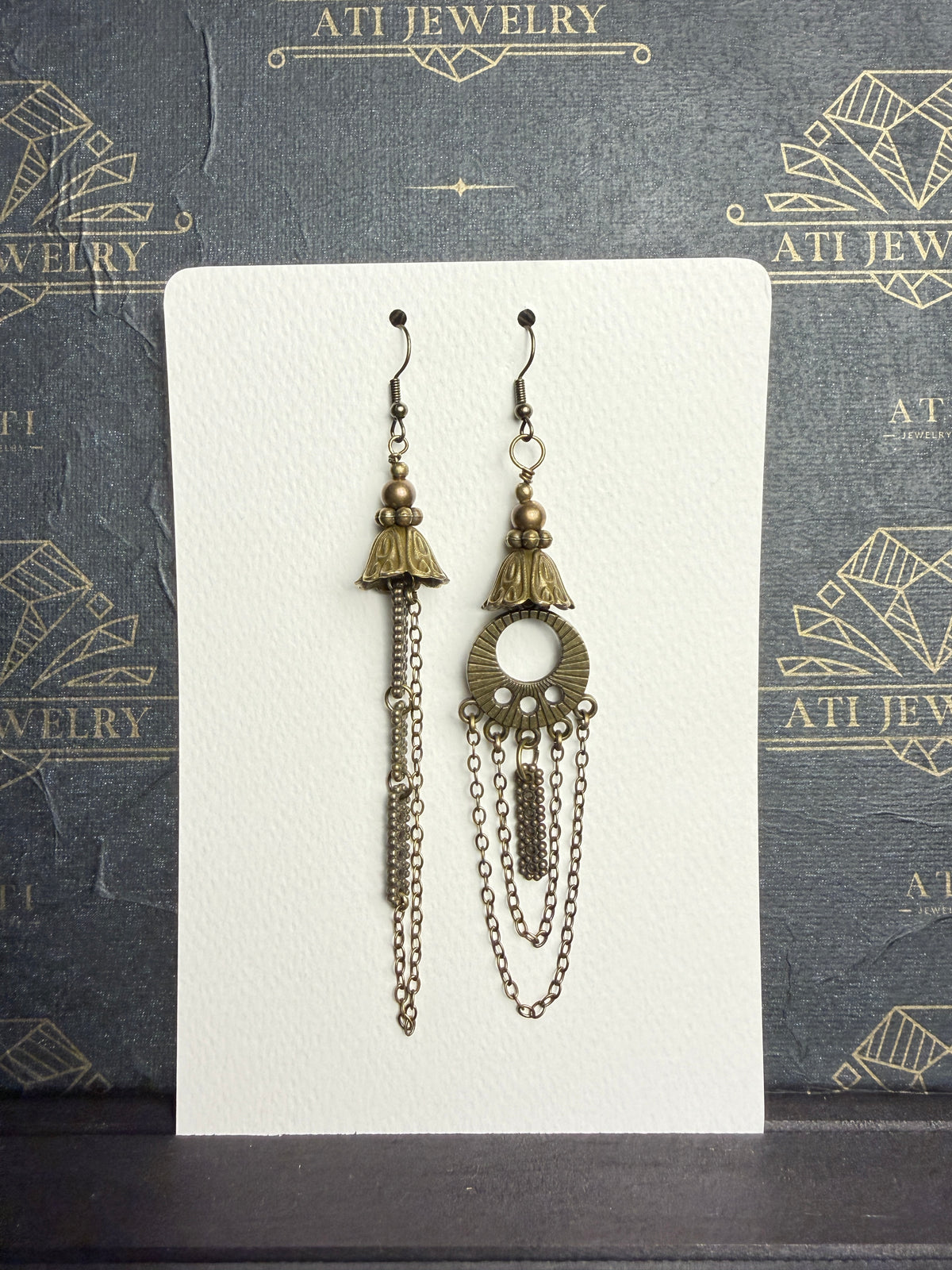 Mismatched antique brass-tone earrings with bell-shaped floral details, long chains, and an open circular ornament on one earring, on an ATI JEWELRY card.
