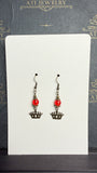 Pair of drop earrings with translucent red beads, antique brass-tone crown charms, and fishhook ear wires on a cream display card.