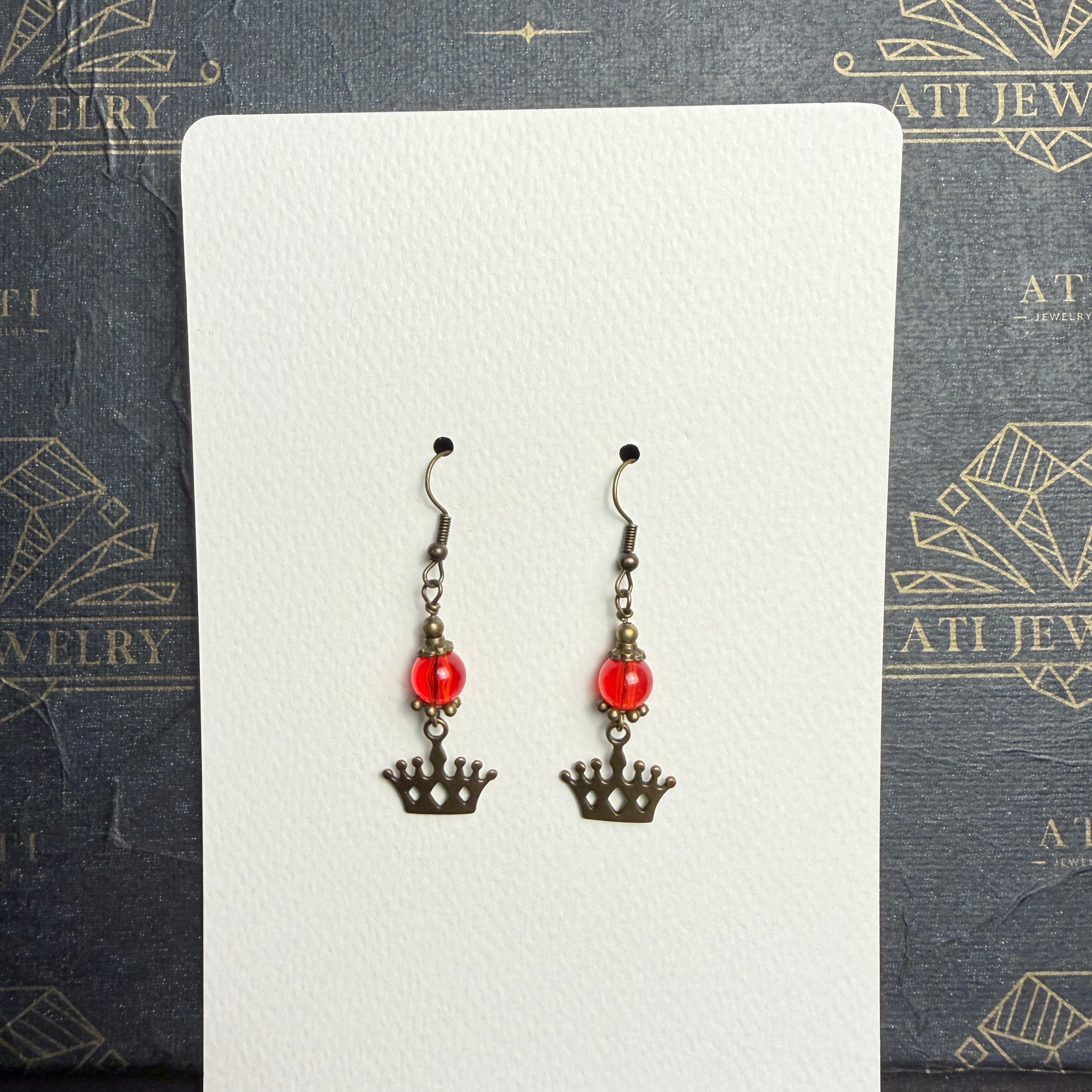 Pair of drop earrings with translucent red beads, antique brass-tone crown charms, and fishhook ear wires on a cream display card.