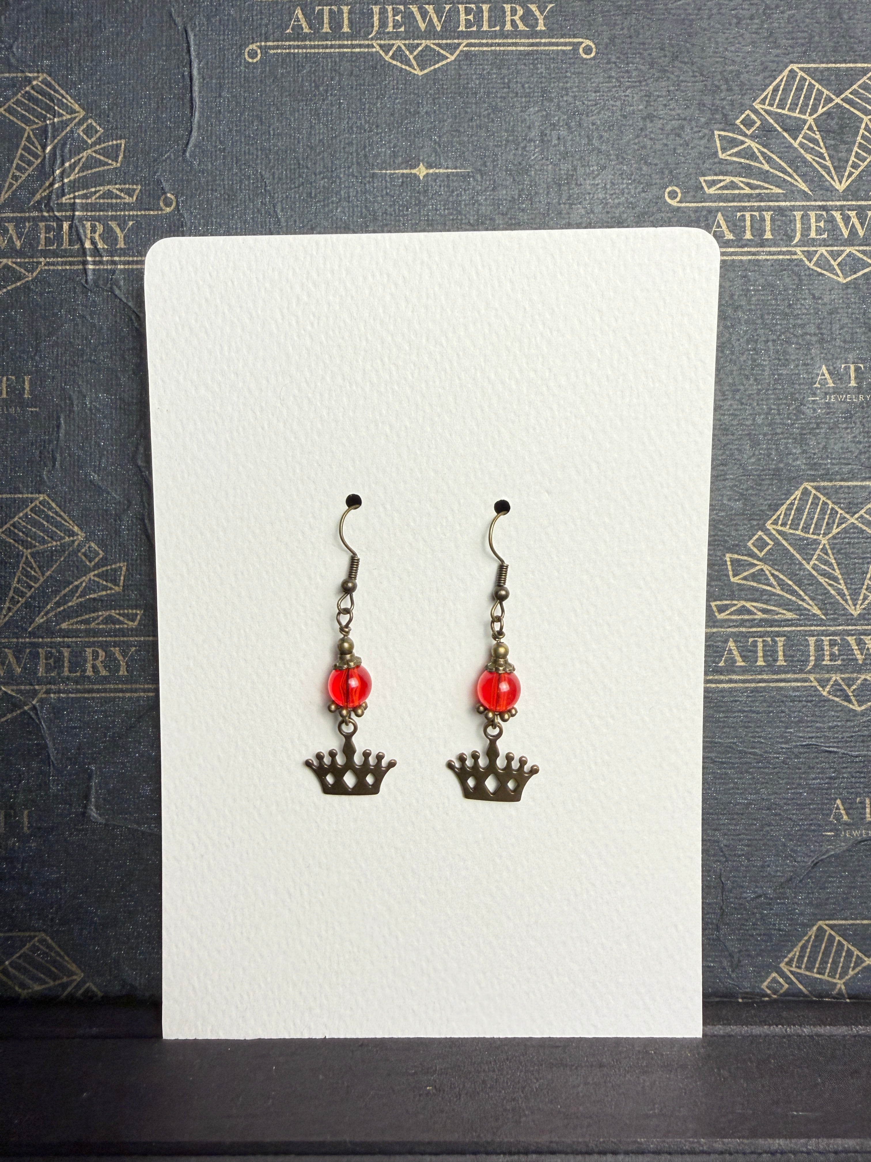Pair of drop earrings with translucent red beads, antique brass-tone crown charms, and fishhook ear wires on a cream display card.